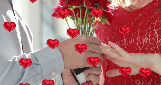 Image of multiple red heart balloons floating over man holding red roses and putting ring on woman f