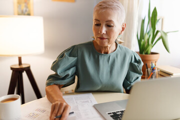 Horizontal indoor portrait of elegant elderly female teacher with stylish haircut working at table in her cabinet in front of laptop, making plan of online lesson, examining papers, typing on keyboard