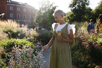 Outdoor side view image of beautiful romantic senior female with stylish look, wearing fashionable dress, walking in park, touching flowers and plants while eating tasty ice cream in waffle cone - 785197604