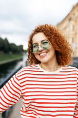 Happy cheerful millennial girl with curly red hair in blue sunglasses and striped sweatshirt walking along big city canal, visiting popular historical places, admiring picturesque cityscape