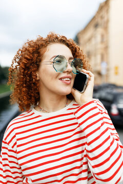 Millennial trendy female hipster with red curly hair hurrying to date, talking on phone with her boyfriend trying to find him on city streets in place of their meeting, wearing funny odd goggles