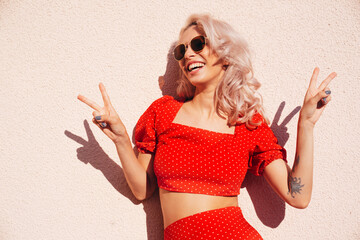 Young beautiful smiling hipster woman in trendy summer red top and skirt clothes. Carefree female posing in street. Positive model near wall. Cheerful and happy. In sunglasses. Shows peace sign