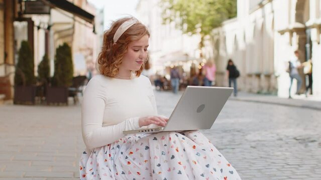 Caucasian woman freelancer sitting on city street using laptop working online distant job browsing website chatting outdoors during break. Girl looks at notebook screen sends messages, watching movies