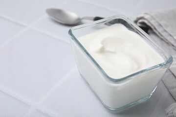 Delicious natural yogurt in glass bowl and spoon on white tiled table, closeup. Space for text