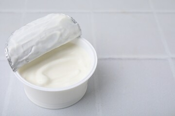 Delicious natural yogurt in plastic cup on white tiled table, closeup. Space for text