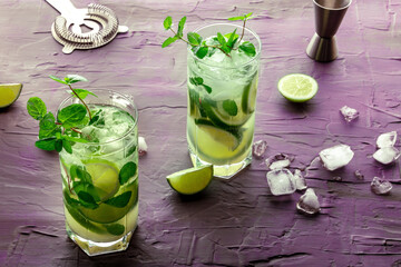 Mojito cocktail, two glasses. Summer cold drink with lime, fresh mint, and ice. Cool beverage on a purple background