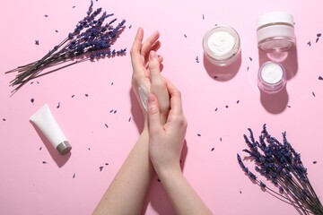 Woman applying hand cream and lavender flowers on pink background, top view