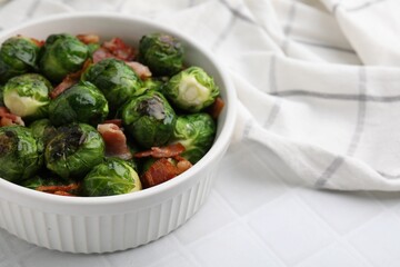 Delicious roasted Brussels sprouts and bacon in bowl on white tiled table, closeup. Space for text