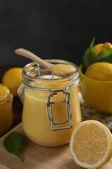 Delicious lemon curd in glass jar, spoon, fresh citrus fruits and green leaves on table