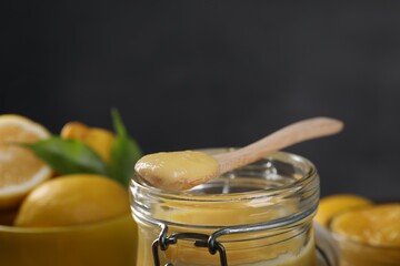 Delicious lemon curd in glass jar, spoon and fresh citrus fruits against blurred background, closeup