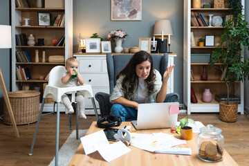 Young mother, business woman having video call on laptop from home while taking care of her baby sitting in tall baby chair. Female business owner on maternity leave must work online from home.