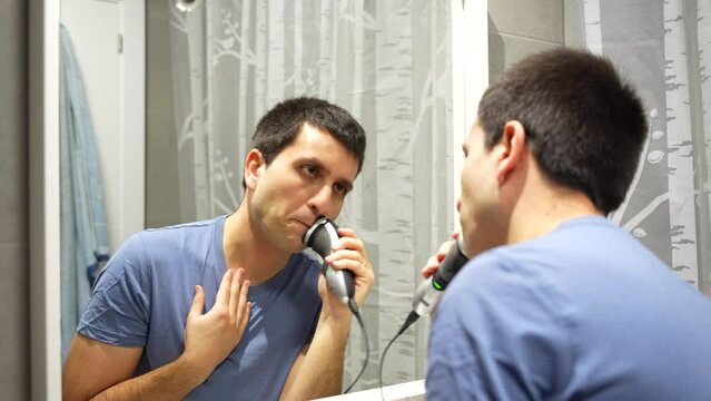 Caucasian man with blue shirt shaving facial hair in bathroom mirror, rear view