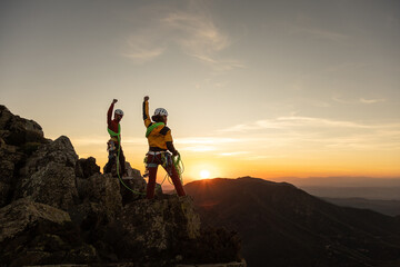 Two people are standing on a mountain top, one of them is holding a fist up in the air. The sun is...