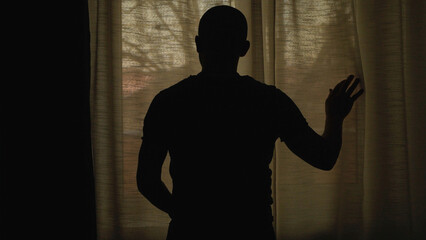Back view of alone man silhouette staring at the window closed with curtains in bedroom.