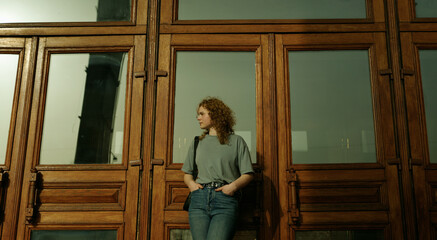 Pensive beautiful young woman with curly hair in grey t-shirt looking away thinking about destiny leaning on wooden doors with windows. Short hairstyle.