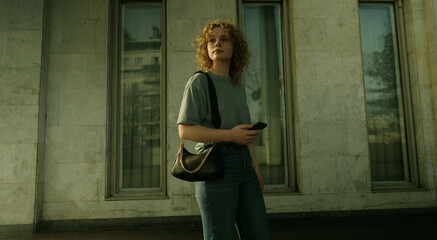 Confident young caucasian woman with curly hair standing in the city looking away holding mobile phone. Woman in casual outfit posing near part of old building.