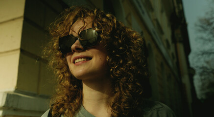 Portrait of attractive smiling woman wearing sunglasses with curly hair walking in urban city street.
