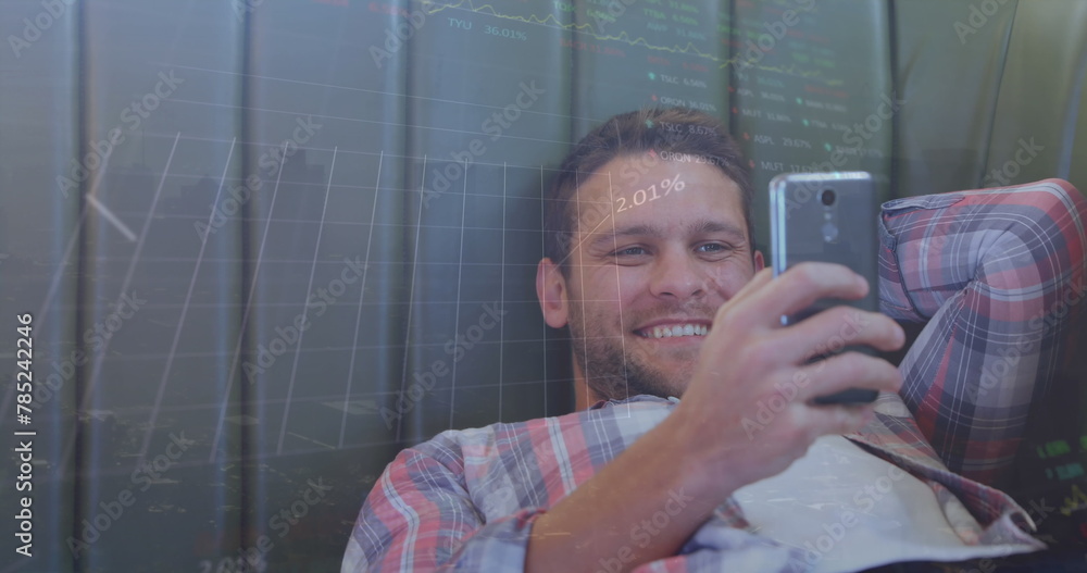 Poster image of graphs and trading boards over smiling caucasian man lying and scrolling on cellphone