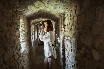 Interested female tourist takes pictures on photo camera while standing in stone corridor of...