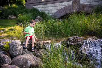 A young boy sits on the river bank, dreams. Warm summer or spring day. Cute Caucasian kid play with water