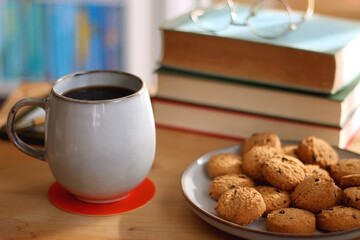 Cup of tea or coffee, pile of books, plate of cookies, reading glasses, e-reader and pen on the table. Colorful rainbow bookshelf in the background. Selective focus.