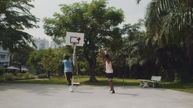 Wide shot of male athlete doing alley-oop with ball while playing streetball with friend on outdoor playground