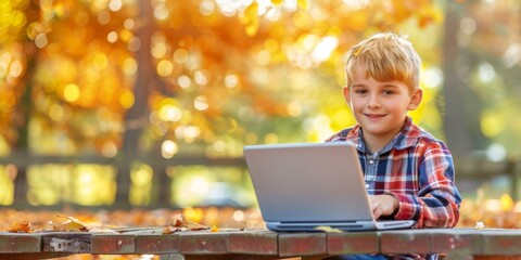 A cheerful young boy uses a laptop on a sunlit park bench surrounded by fall foliage.