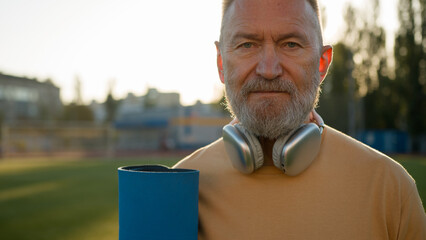 Old Caucasian man pensioner looking around headphones holding mat sport stadium outside strong...