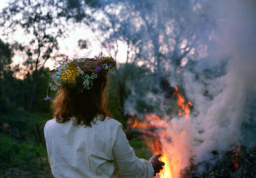 Woman in floral wreath near campfire in forest, dark nature background. rear view. Magic ceremonial, witchcraft. witch wiccan ritual for Beltane, Midsummer, Litha.