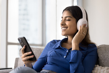 Positive relaxed young 20s Indian girl in big headphones listening to calm tranquil music from online playlist on smartphone, touching earphones, smiling, resting on home sofa, enjoying leisure