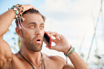 Handsome shirtless tanned man shows surprised expression while talking on the phone on the promenade on a summer day.