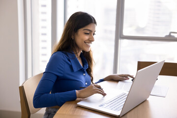 Happy focused Indian freelancer woman working at laptop at home, sitting at workplace table, using freelance job application, modern technology for business Internet communication