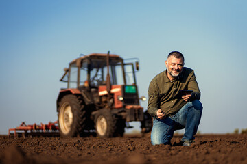 Portrait of satisfied mature farmer standing in field with tablet supervises the cultivation of soil. - obrazy, fototapety, plakaty