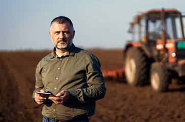 Portrait of satisfied mature farmer standing in field with tablet supervises the cultivation of soil.