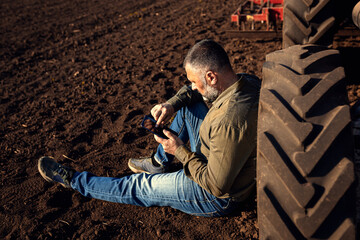 Farmer siting and using tablet while resting after cultivation of soil leaning on tractor tire.