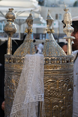 A white scarf with tassels gently draped over a silver case enclosing a Sefardi style Torah scroll.