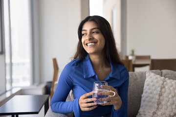 Happy attractive young 20s Indian girl holding glass, refreshing, drinking clear fresh water, looking away with toothy smile, enjoying healthy nutrition, keeping diet, aqua balance