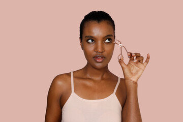 Jung stunning African American woman wearing pink sleeveless top is using eyelashes curler. Make-up...