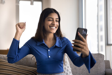 Joyful excited young Indian winner woman enjoying online victory, making hand yes gesture, looking at mobile phone, smiling, using Internet application on telephone for success