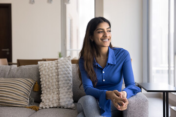 Cheerful dreamy beautiful Indian woman sitting on comfortable couch, enjoying leisure in cozy apartment, looking at window away, smiling, thinking, daydreaming, planning happy future