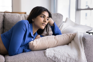 Thoughtful attractive 20s Indian girl resting on comfortable home couch, leaning head on hand,...