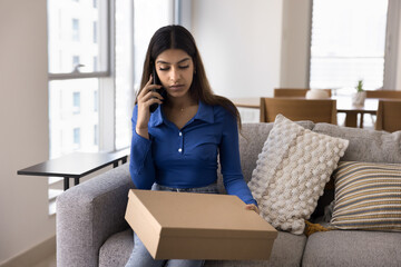 Serious young Asian consumer woman holding cardboard box with purchase from Internet store, making telephone call for returning rejected goods, talking on mobile phone over parcel