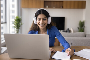 Cheerful attractive young Indian student girl in big white headphones talking to teacher on video call, watching online class, conference, smiling, writing notes, studying from home