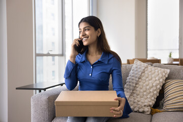 Cheerful satisfied young Indian consumer woman holding paper cardboard box with purchase from Internet shop, webstore, speaking on mobile phone, smiling, ordering delivery shipping service for sending