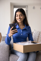 Happy 20s Indian girl using ecommerce application on smartphone, holding open cardboard box with purchase from online store inside, giving positive good feedback, smiling. Vertical shot
