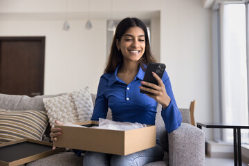 Cheerful young Indian customer girl typing on smartphone over open logistic cardboard box, unpacking parcel from delivery transportation service, chatting on mobile phone, smiling