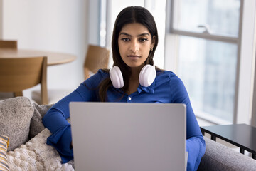 Serious young Indian student girl studying at home, holding laptop on lap, resting on home couch, typing, using big wireless headphones. Freelancer woman working on online communication project