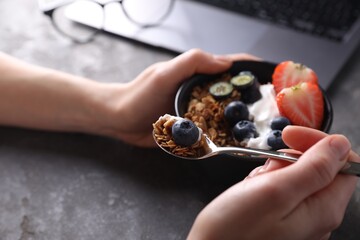 Woman eating tasty granola with yogurt and berries at workplace, closeup
