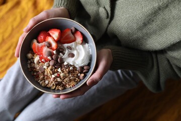 Woman holding bowl of tasty granola with chocolate chips, strawberries and yogurt indoors, top view