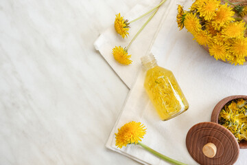 Bottle with cosmetic oil and plate of dandelion flowers on a white marble background. Top view, flat lay. Space for text.
